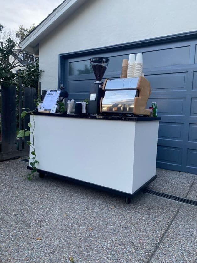 Coffee stand with espresso machine and supplies outside a residential garage.